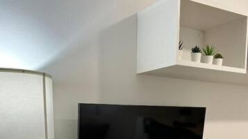 Una sala moderna con una televisión de pantalla plana montada en la pared, una consola blanca con objetos decorativos y un estante blanco con pequeñas plantas.