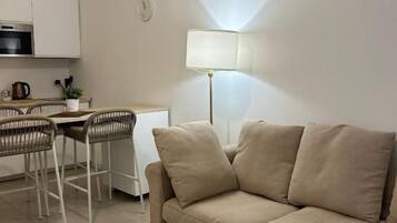 Een woonkamer met een bank, een eettafel met stoelen, een lamp en een keuken met kasten en apparatuur.