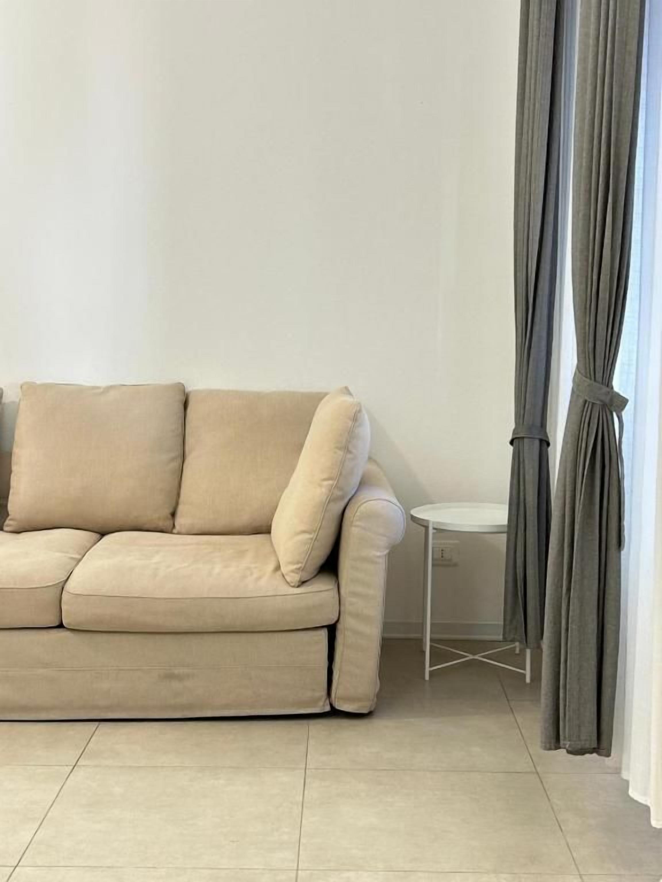 Un canapé beige, une petite table ronde et des rideaux gris dans une pièce dont le mur est d’un blanc uni et le sol carrelé.