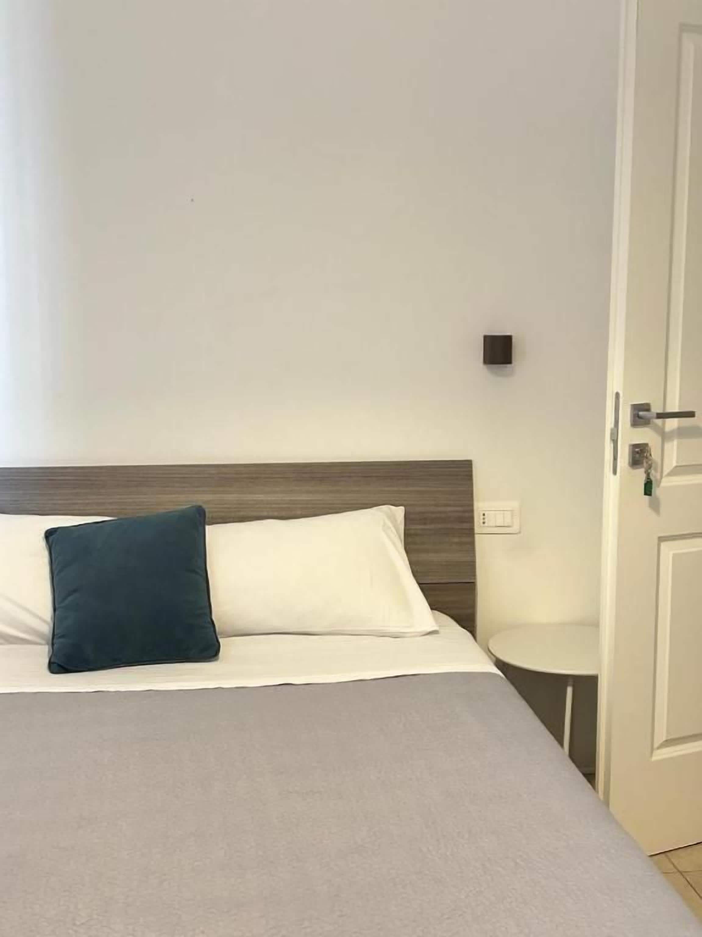 A bed with a white headboard and a dark pillow, a small bedside table, and a door to the right.
