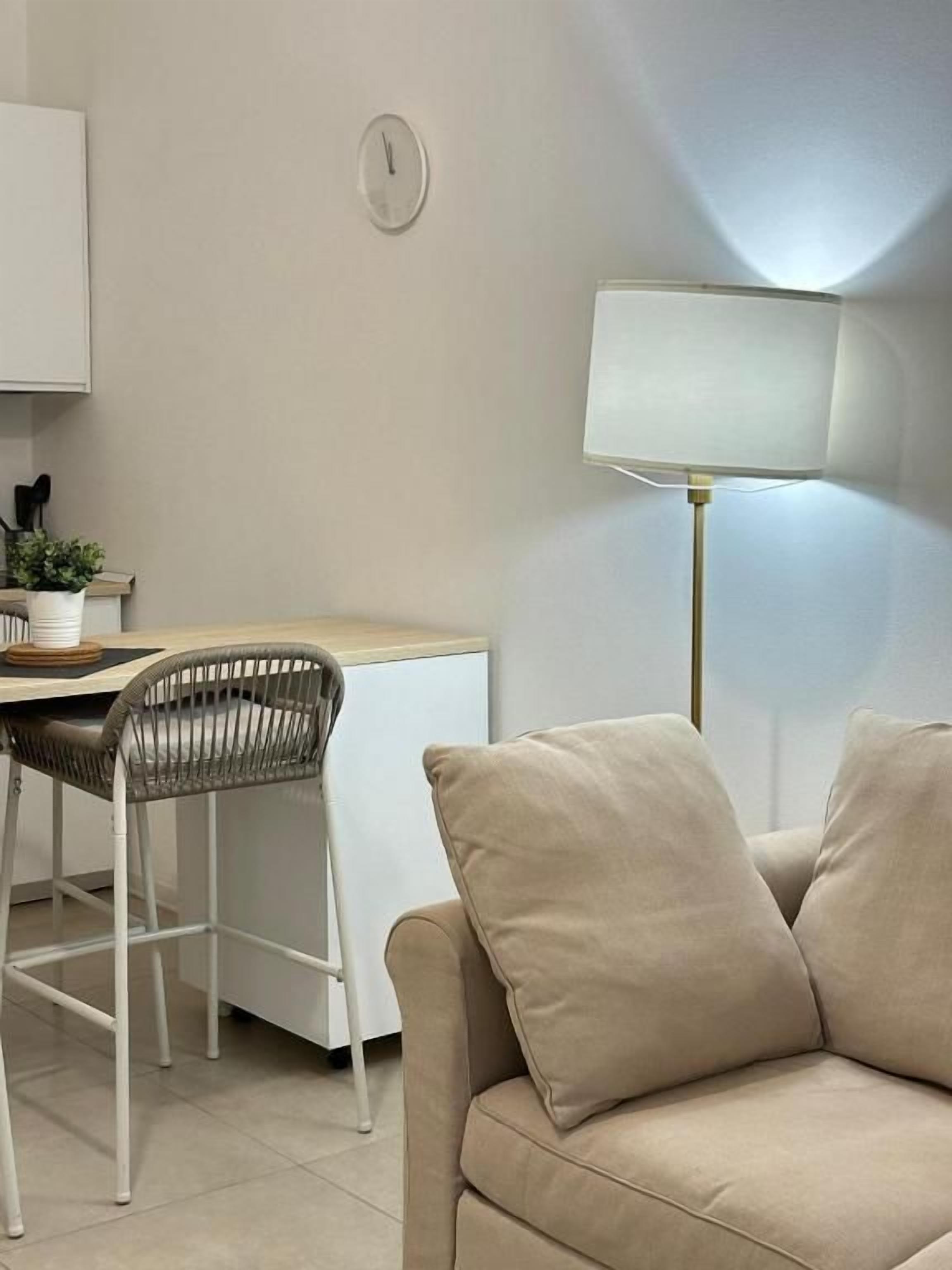 A modern living room with a sofa, a dining table with chairs, a lamp, and a clock on the wall.