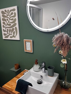 Une salle de bain avec un lavabo blanc, un miroir avec un éclairage intégré, un tableau encadré et un vase avec des fleurs.