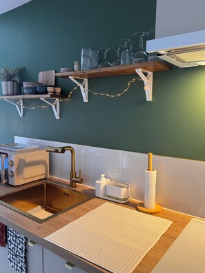 Une cuisine équipée d’un four à micro-ondes, d’un évier et d’un plan de travail en bois. Des étagères au-dessus du plan de travail servent à ranger de la vaisselle et une plante.