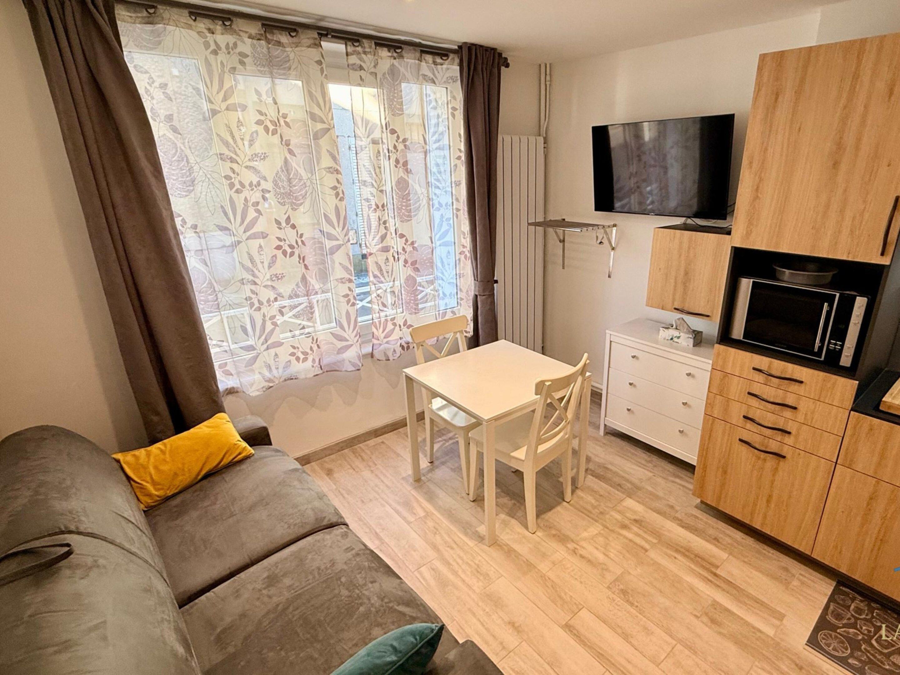A compact living space with a sofa, a dining table, a TV mounted on the wall, and a kitchenette with a microwave.
