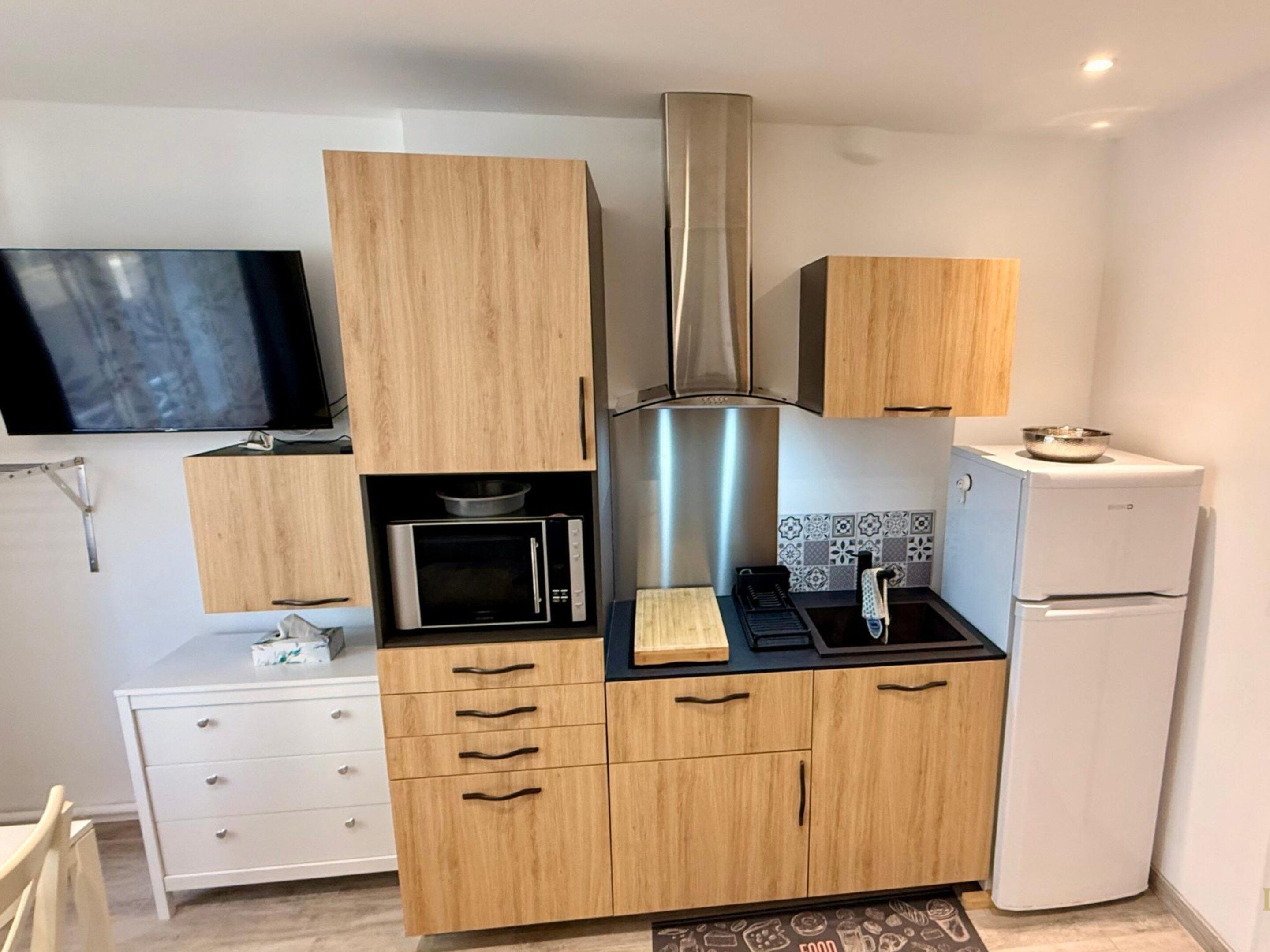 A compact kitchen with wooden cabinets, a microwave, a stainless steel range hood, a white refrigerator, and a flat-screen TV mounted on the wall.