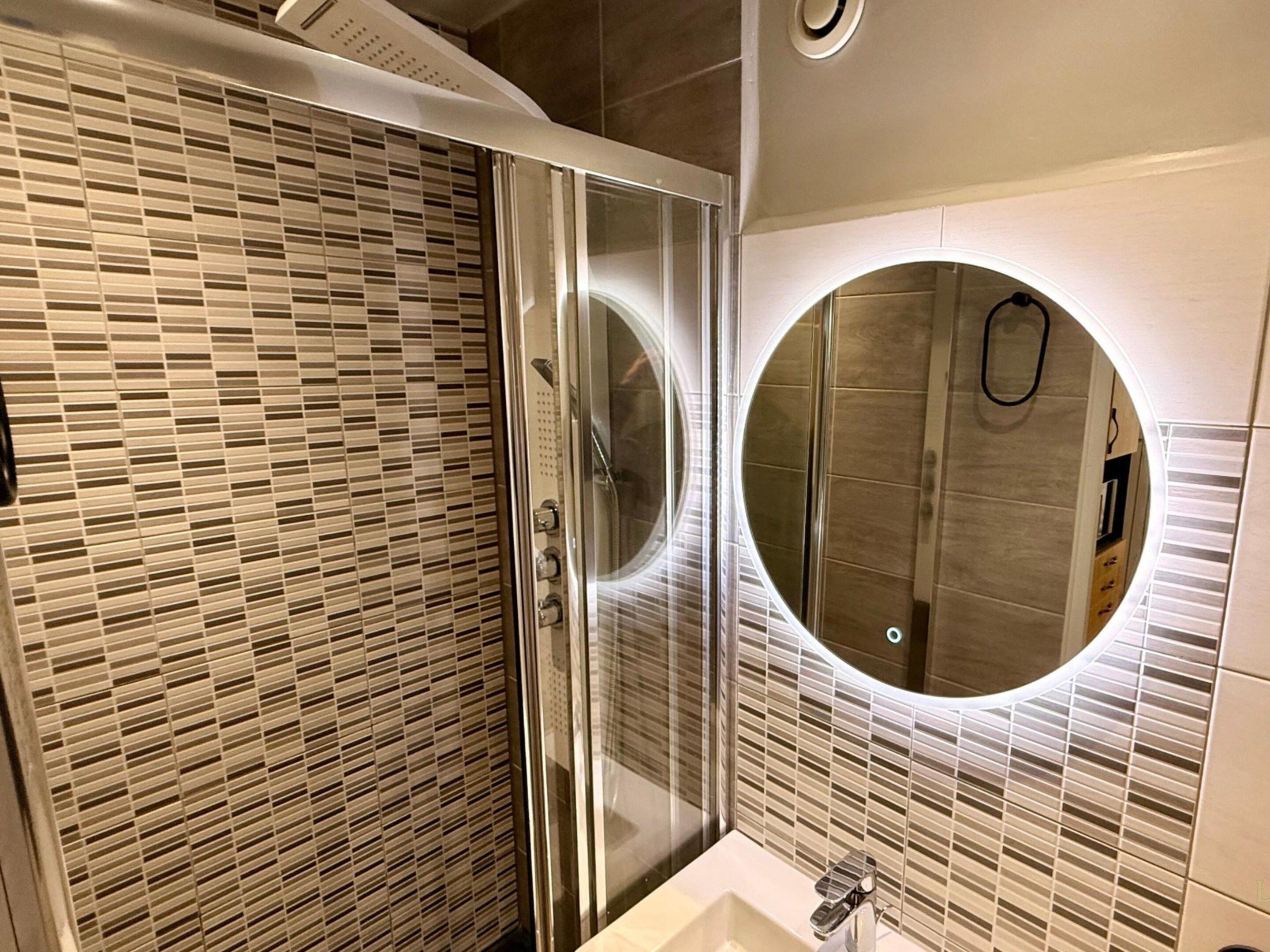 A modern bathroom with a glass shower enclosure, a round mirror, and a tiled wall.