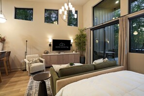 Habitación de hotel moderna con una cama grande, un sofá, un escritorio con televisor de pantalla plana y un ventanal con vistas a árboles.