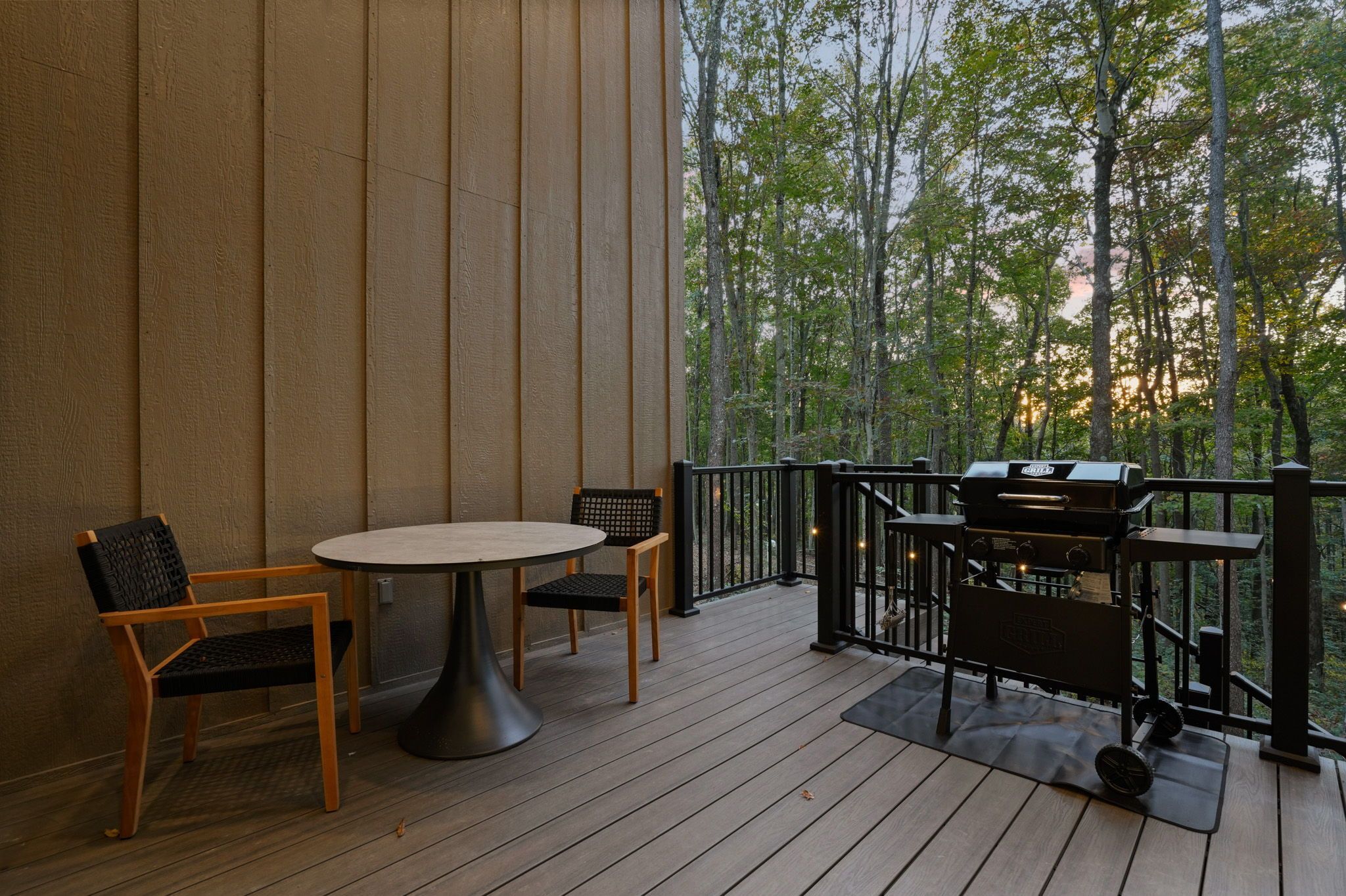 Terraza exterior con una mesa redonda y dos sillas, una parrilla y vistas a árboles.