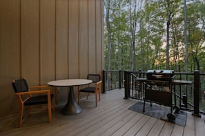 Terraza exterior con una mesa redonda y dos sillas, una parrilla y vistas a árboles.