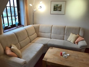 A corner sectional sofa with cushions, a wooden coffee table with a candle and a glass, and a painting on the wall. - Hansen Nieblum - Holiday Apartment 1 (Nieblum)