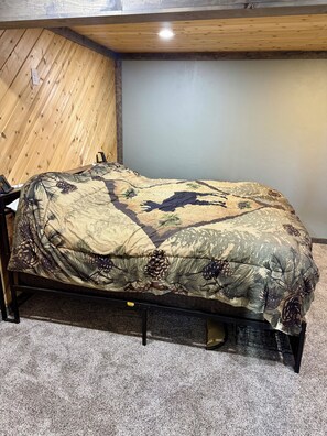 Ein Einzelbett mit einem zerrissenen Tarnmuster, in einem Zimmer mit Holzwänden und Teppichboden.