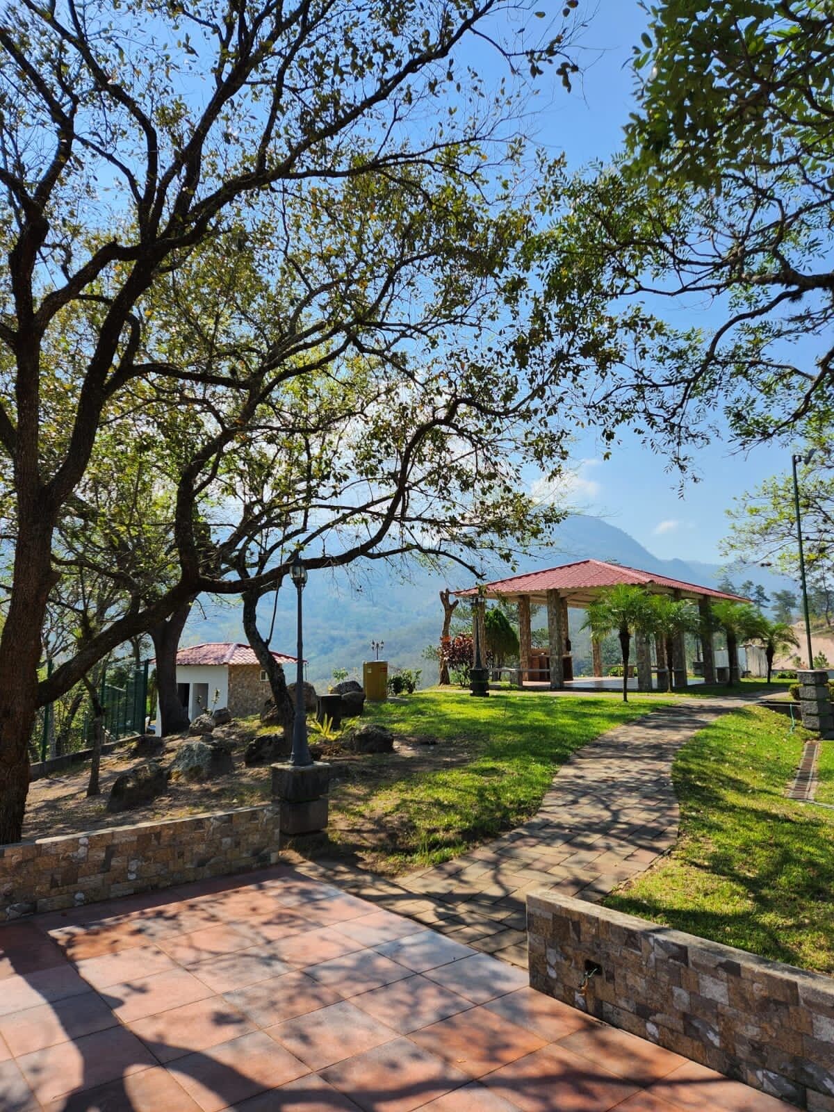 A gazebo with a red roof, surrounded by a stone wall and a paved walkway.
