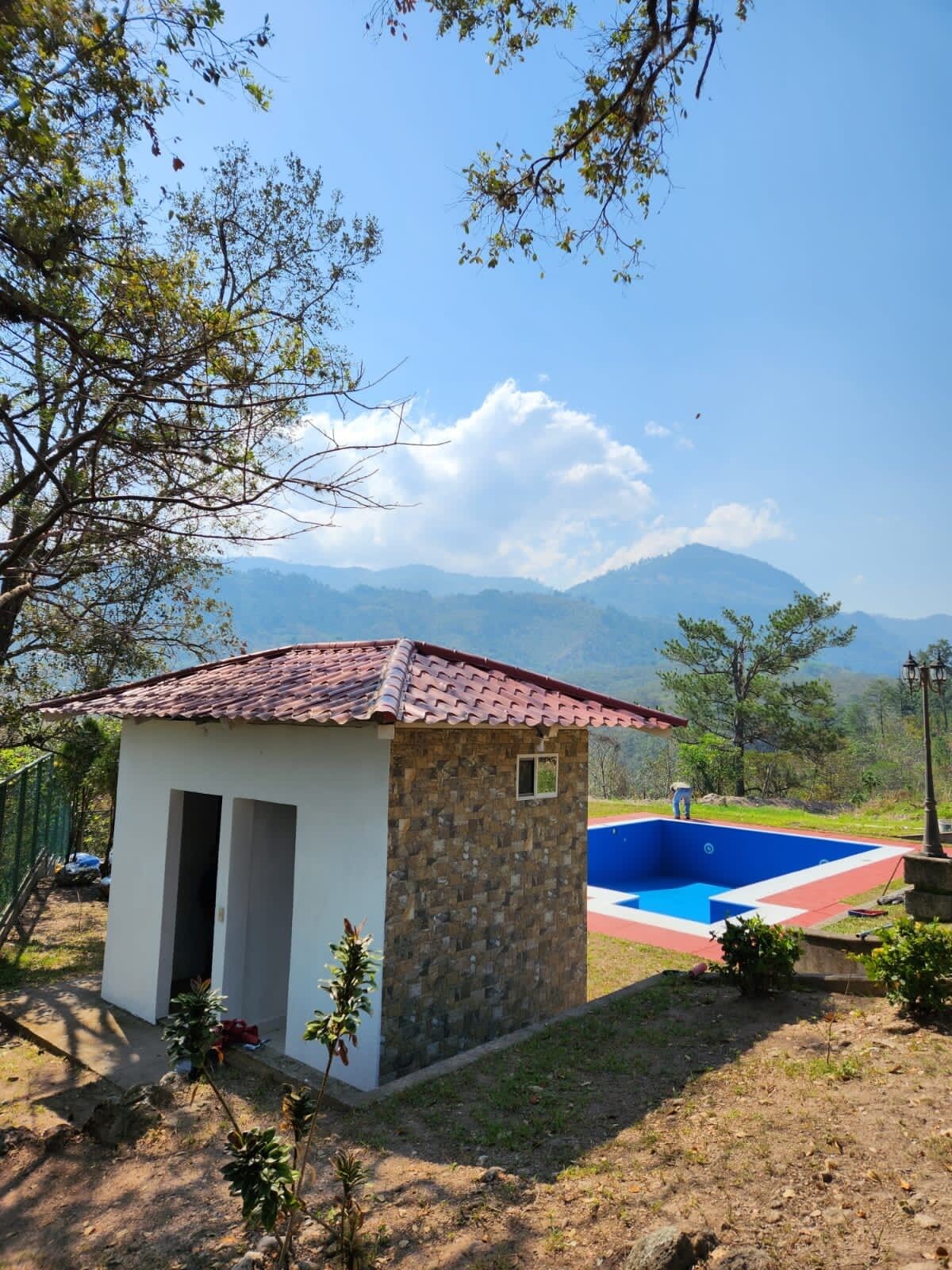 A small stone building with a tiled roof, a swimming pool, and a view of distant mountains.