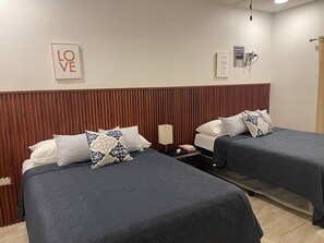 A hotel room with two beds, each with a dark bedspread and patterned pillows. There are two wall-mounted light fixtures and two framed inspirational quotes.