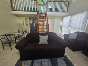 Een woonkamer met een bank, twee fauteuils, een eettafel met stoelen, een keukenhoek en een trap naar een verdieping.