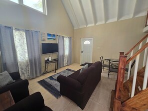Een woonkamer met een donkerbruine bank, een flatscreen televisie aan de muur, een eettafel met stoelen en een houten trap.