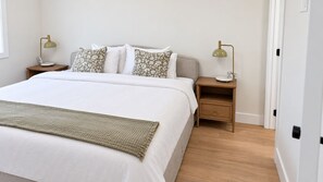 Un lit bien fait, recouvert d’une couette blanche et d’un plaid vert, avec deux tables de chevet de chaque côté, chacune surmontée d’une lampe, et une petite table de nuit également dotée d’une lampe.