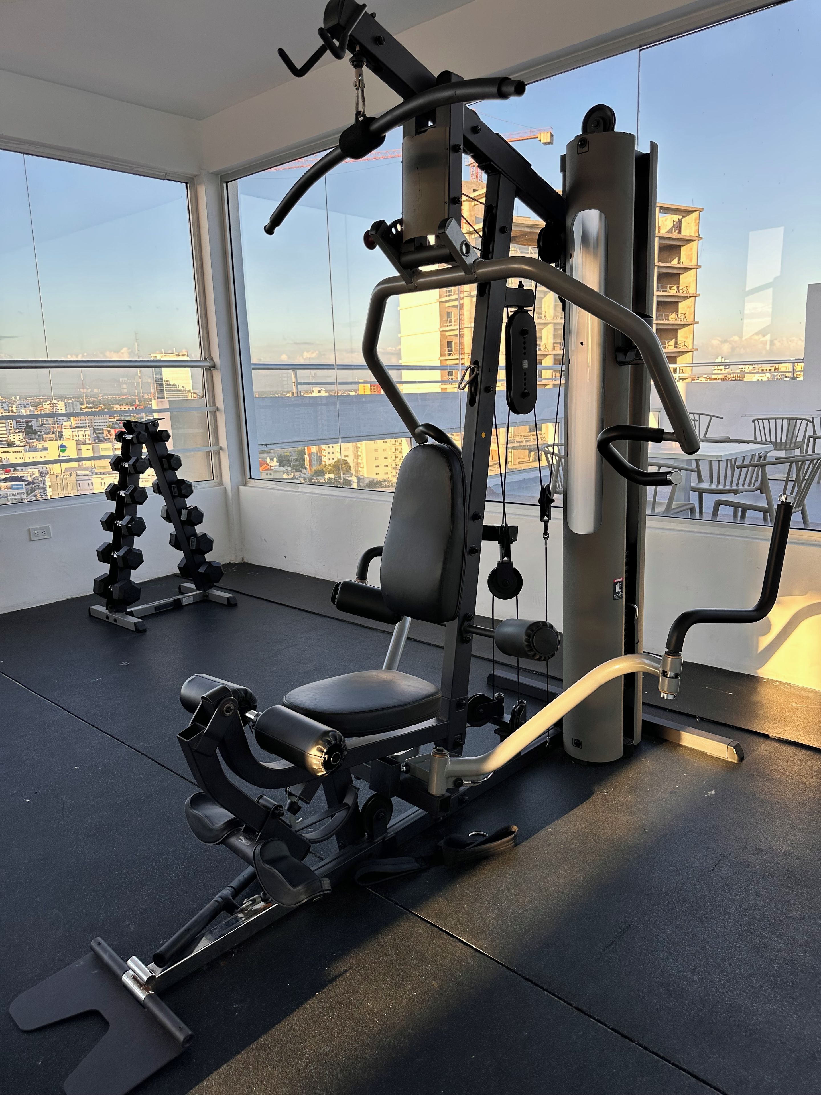 Un gimnasio con equipamiento de ejercicio, una banca y vistas de la ciudad a través de amplios ventanales.