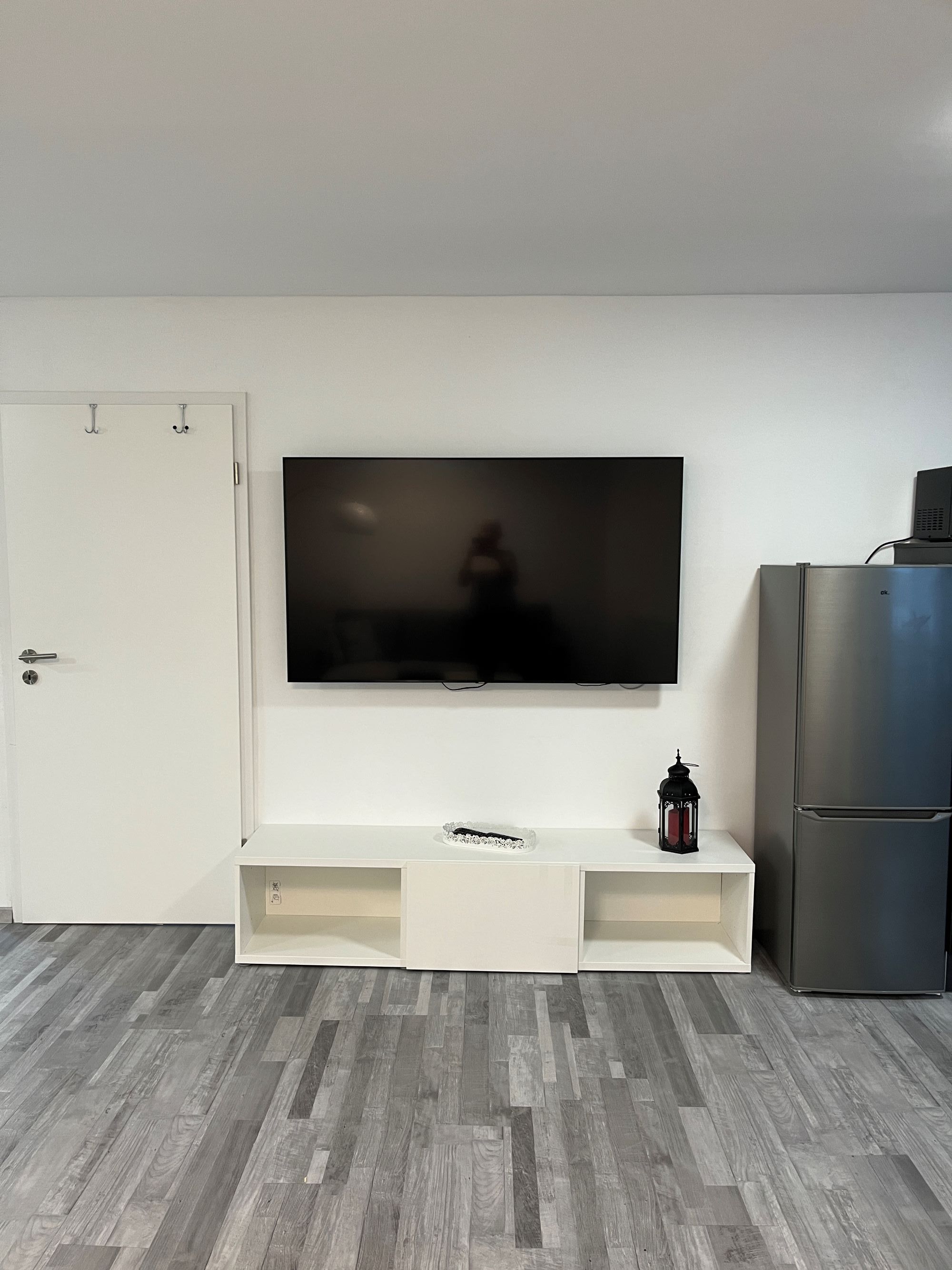 Een woonkamer met een flatscreen televisie aan de muur, een witte entertainmentkast eronder, een koelkast aan de rechterkant en een houten vloer.