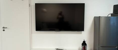 Een woonkamer met een flatscreen televisie aan de muur, een witte entertainmentkast eronder, een koelkast aan de rechterkant en een houten vloer.