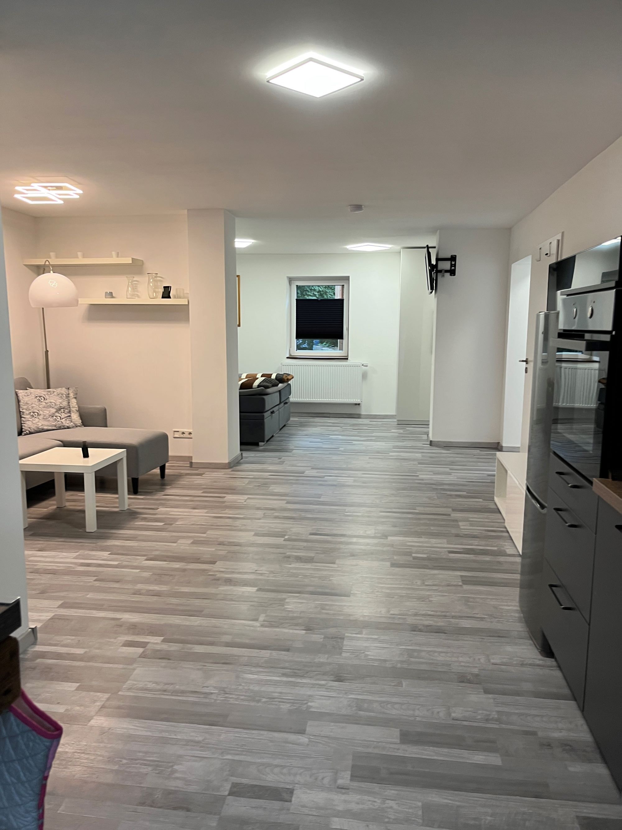 Eine moderne Küche mit einer zentralen Kochinsel, ein Essbereich mit einer Couch und einem Tisch sowie ein Flur zu einem Fenster.