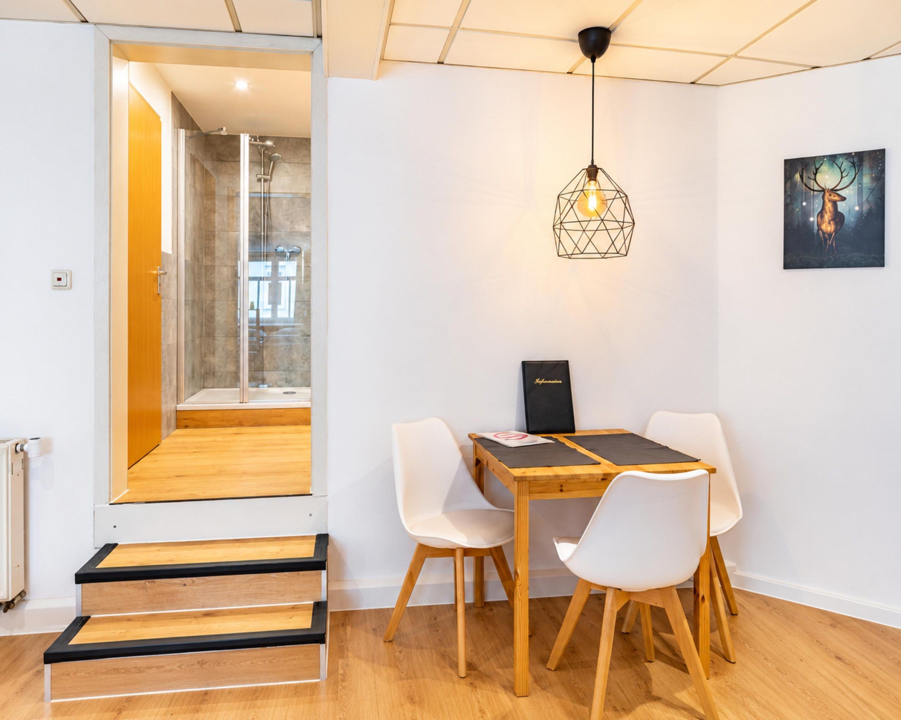 A modern interior with a wooden dining table, white chairs, a geometric pendant light, and a framed artwork on the wall.
