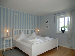 A bedroom with a white bed, striped wallpaper, a window with a plant, and a bedside table with a lamp. - Holiday Home in Oevenum With Swimming Pool (Oevenum)