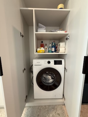 A compact laundry room with a white washing machine, shelves above containing cleaning supplies, and a small storage area. - Cozy clean & minimalistic apartment near Airport, with balcony & view! (Chișinău)