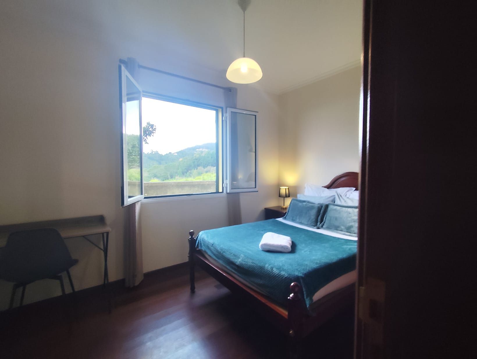 Un dormitorio con cama, mesita de noche, lámpara, sillón y ventana con vistas a zonas verdes.