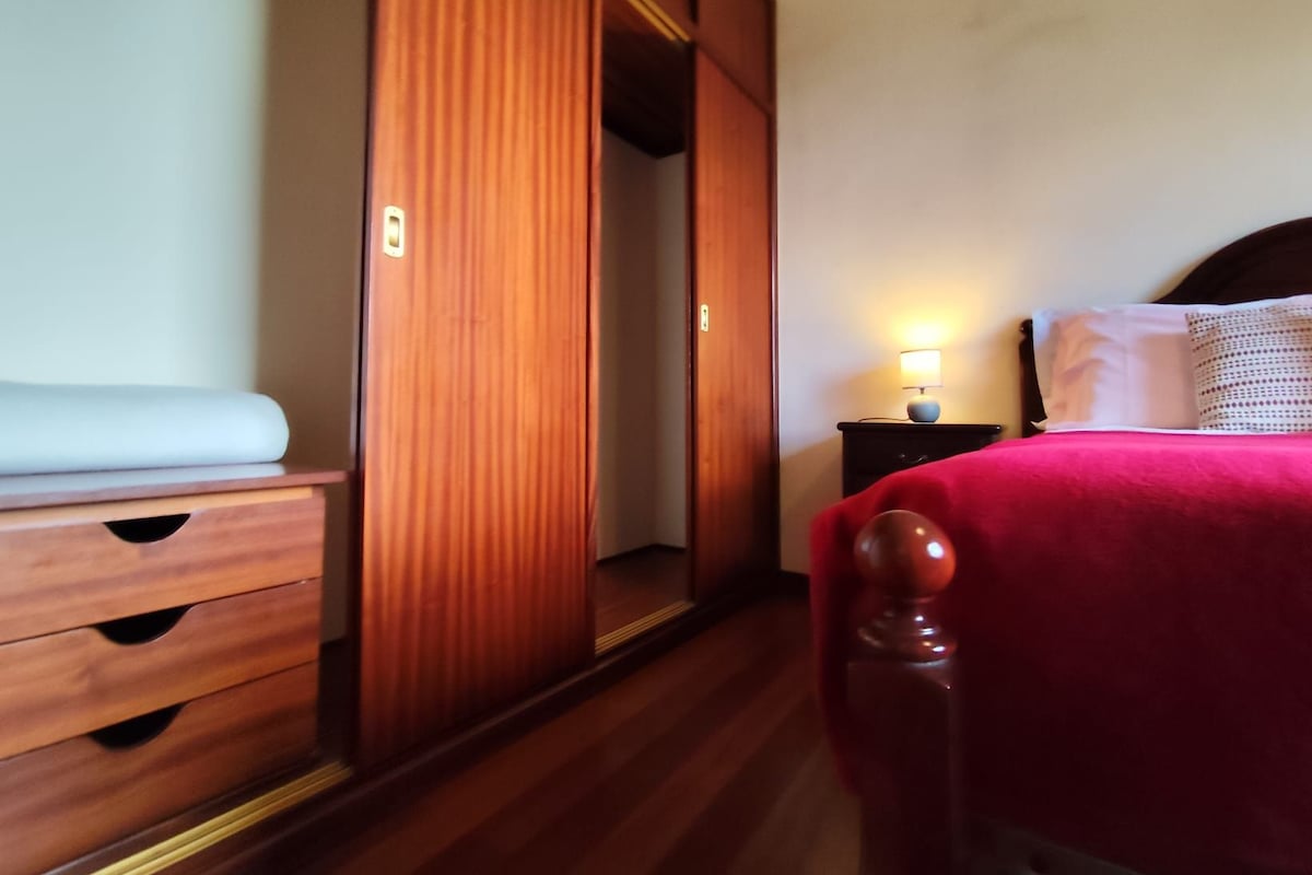 Une chambre d’hôtel avec une armoire en bois, une table de chevet avec une lampe et un lit recouvert d’une couverture rouge.