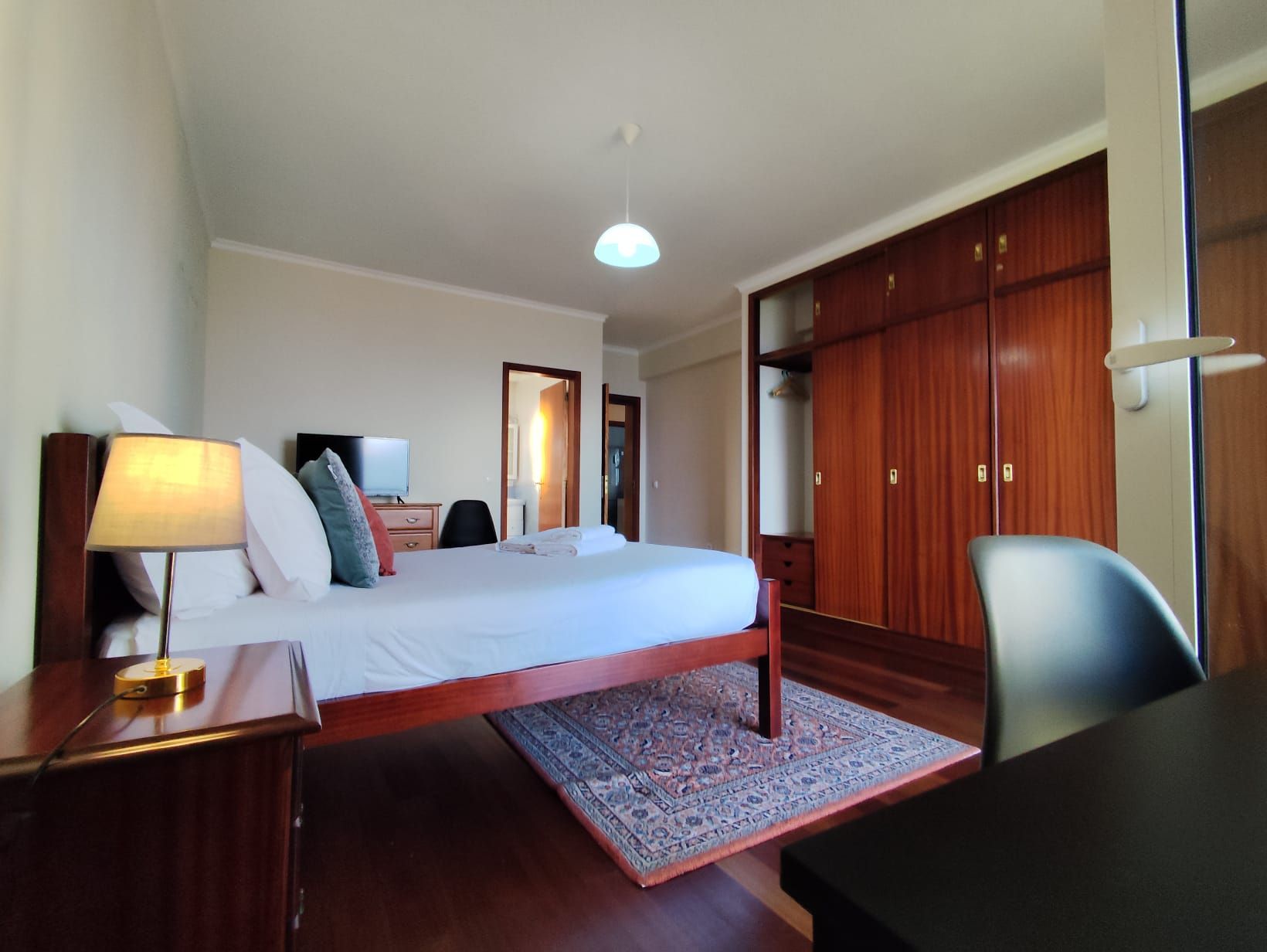 Une chambre d’hôtel comprenant un lit, une table de chevet, une lampe, une télévision, une armoire et un tapis à motifs.