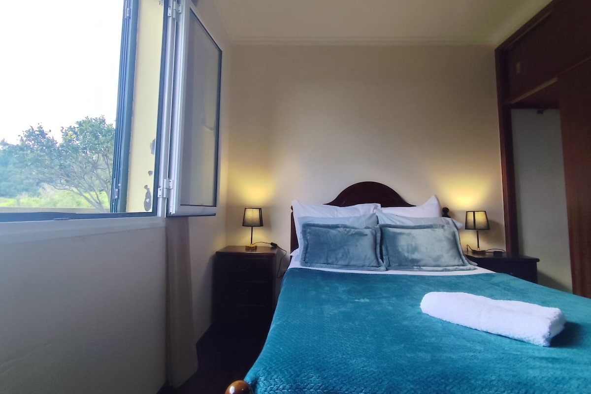 Une chambre à coucher avec un lit, deux lampes de chevet, une fenêtre et une vue sur la verdure.