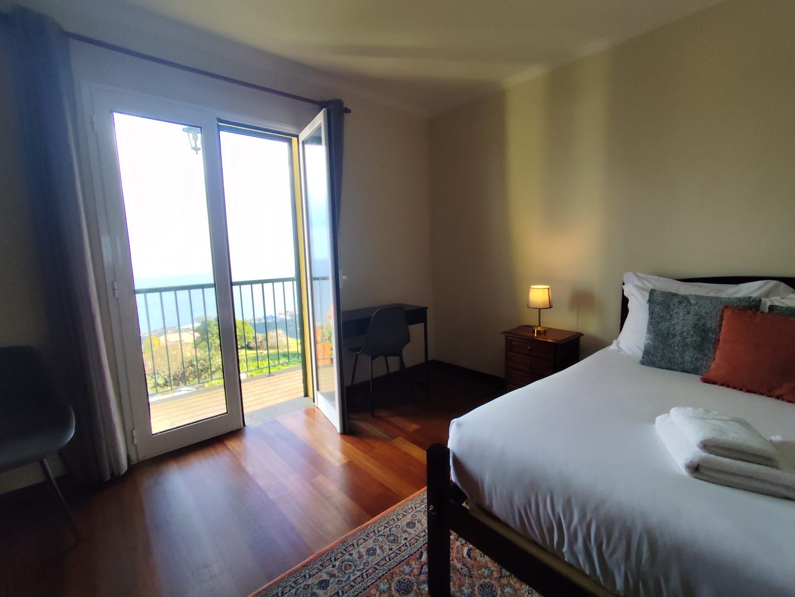 Una habitación de hotel con cama, escritorio, silla, mesita de noche con lámpara y balcón con vistas.