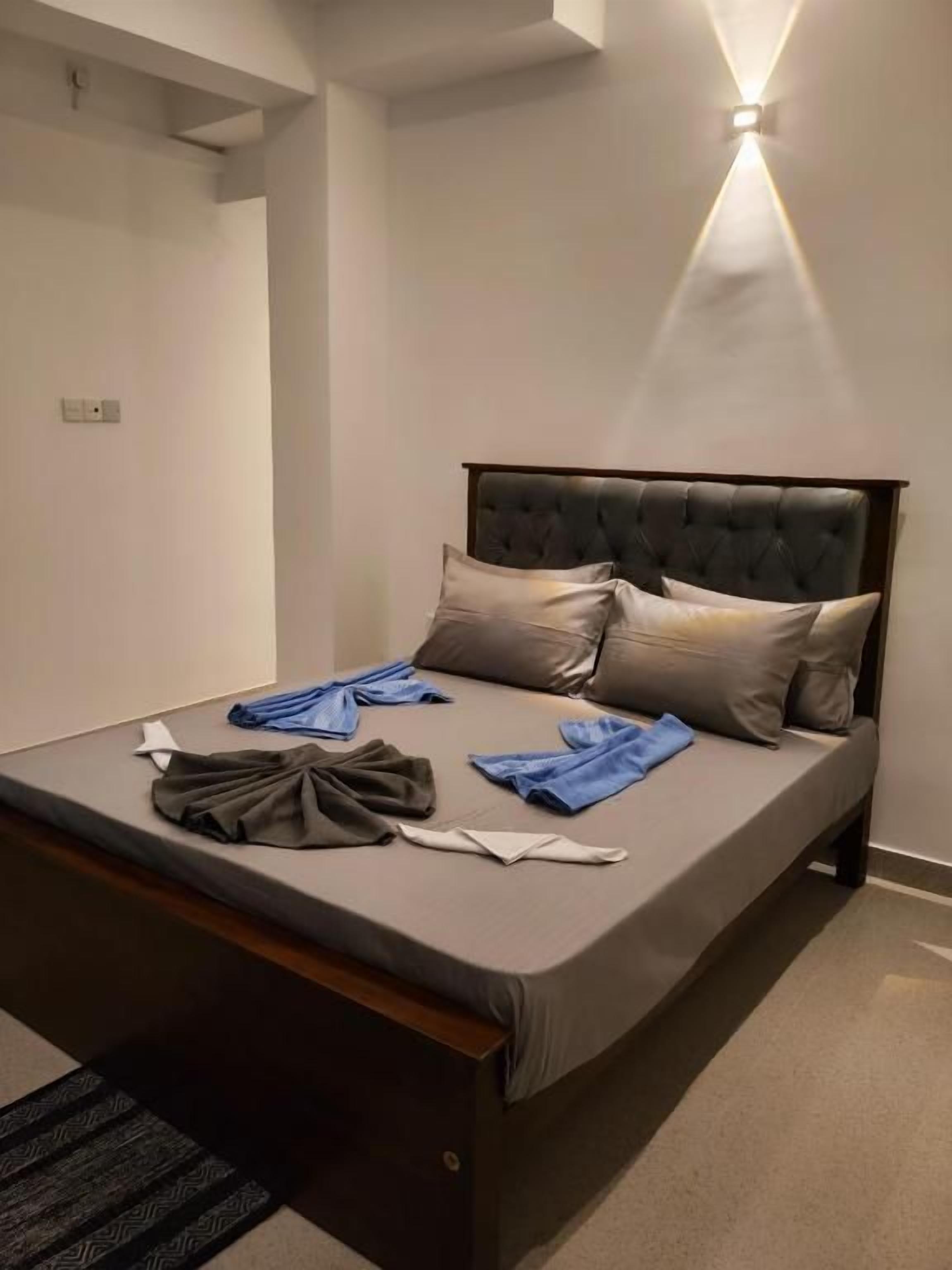A neatly made bed with pillows and folded towels, a dark headboard, and a wall-mounted light fixture.