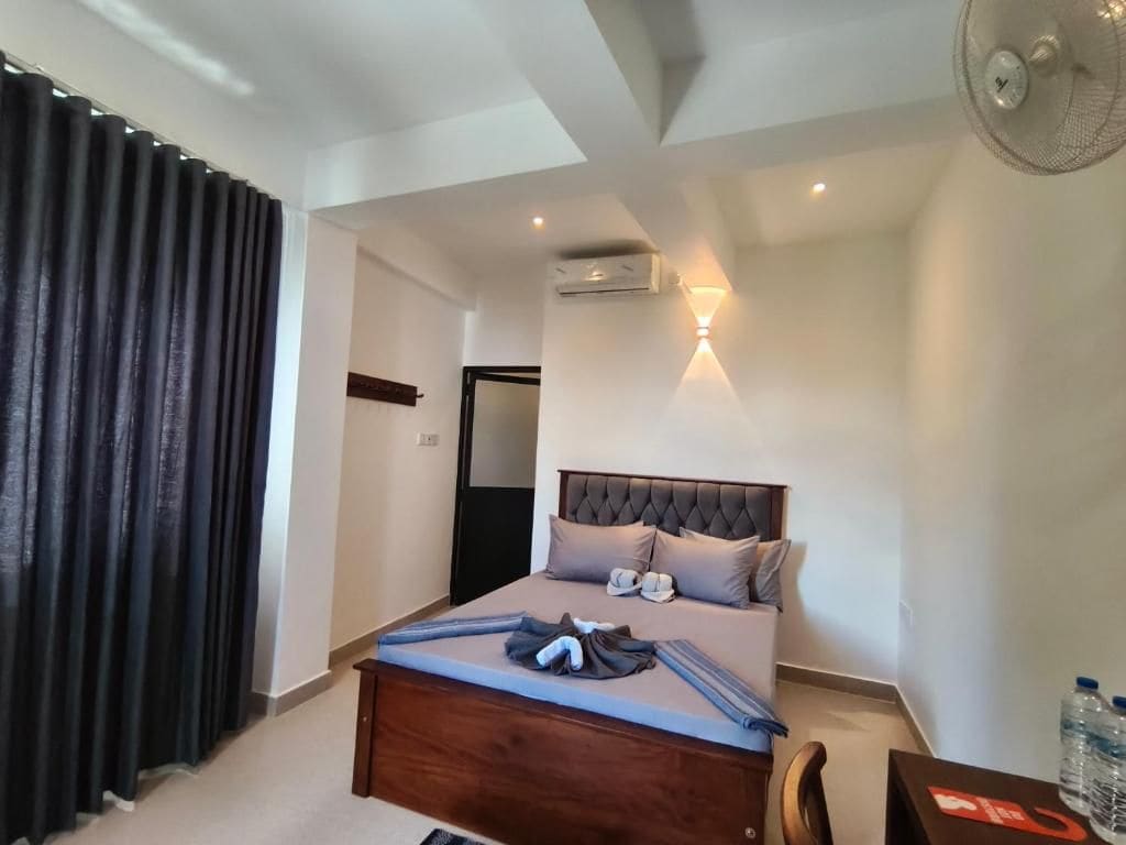 A hotel room with a bed, a desk with water bottles, a ceiling fan, and a wall-mounted light fixture.