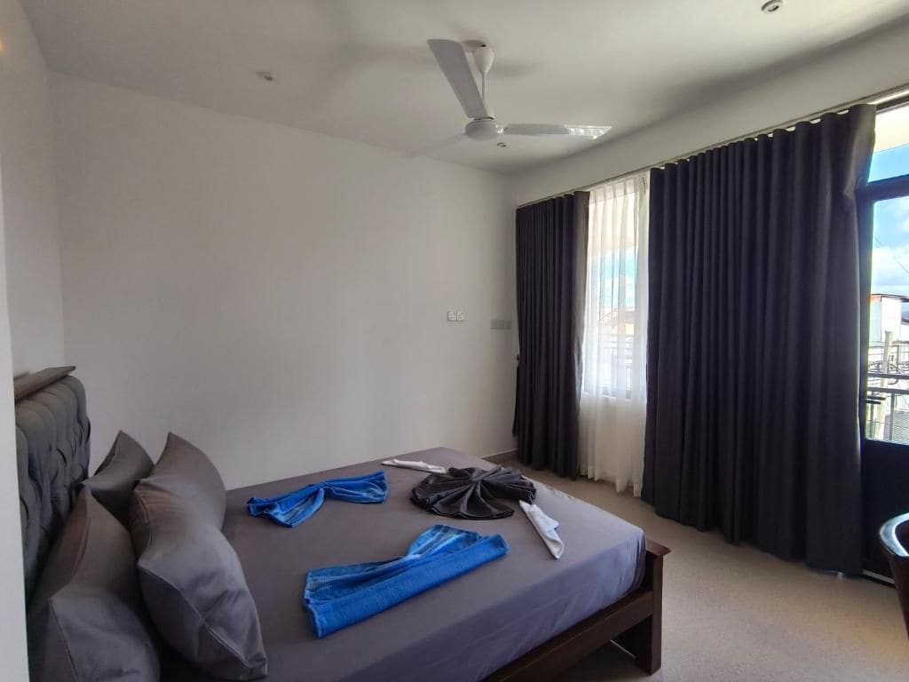 A hotel room with a bed, a ceiling fan, a window with curtains, and a view of buildings outside.
