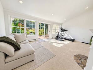 Ett rymligt vardagsrum med en beige soffa, ett löpband och ett stort fönster med utsikt över grönområden.