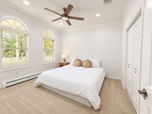 A bedroom with a bed, a ceiling fan, two windows with blinds, a nightstand with a lamp, and a door. - Shirley Mansion - Family Stay, 3BR/2.5BA One-Level, Office, Sunroom, Yard (Shirley)