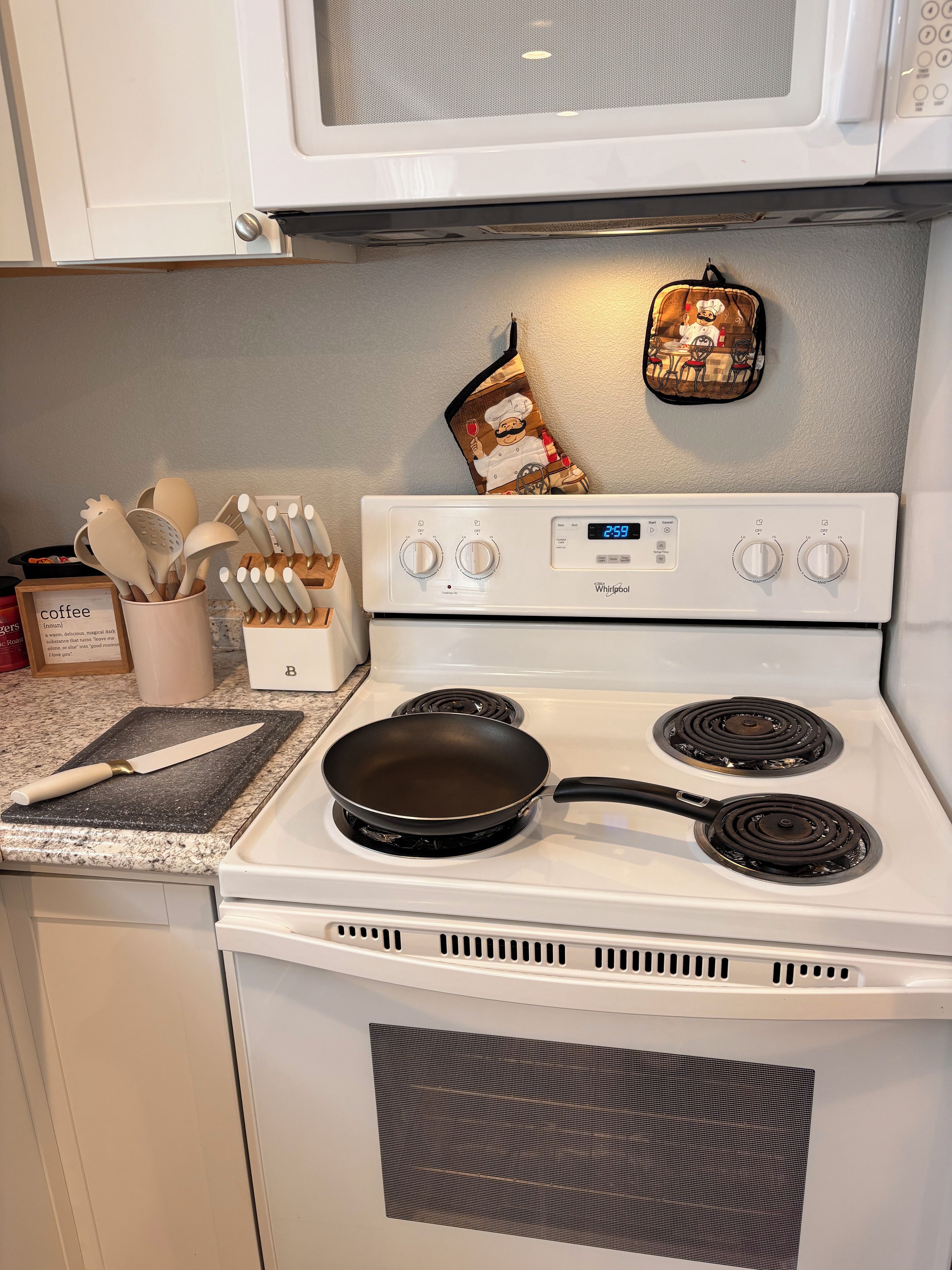 Una cocina con estufa blanca, dos hornillas, un microondas encima y una espátula sobre la encimera.