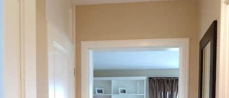 A hallway with a ceiling light fixture, a door on the left, and a view into a room with a closet and curtains.