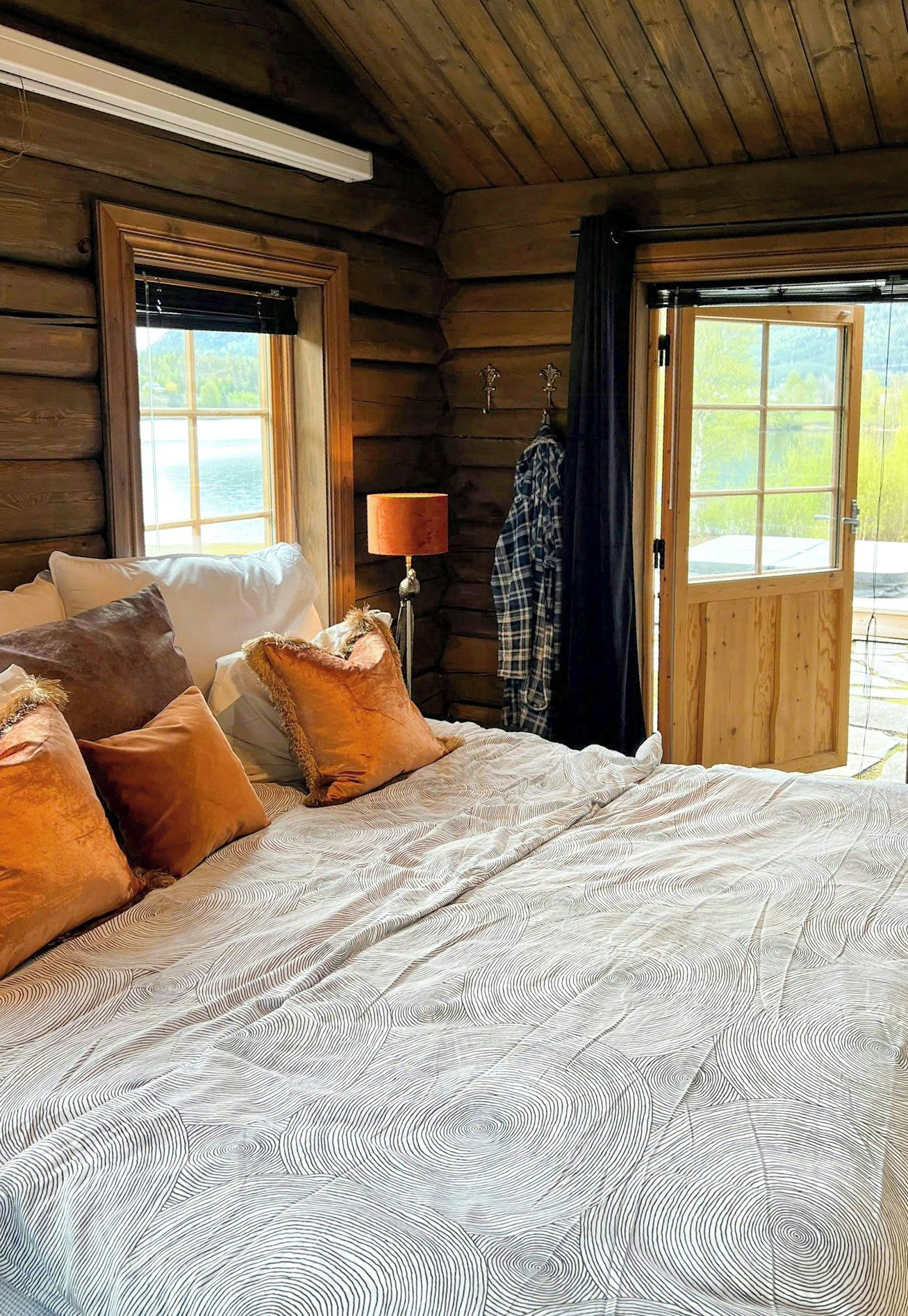 A log cabin bedroom with a bed, pillows, a lamp, and a view of the outdoors.