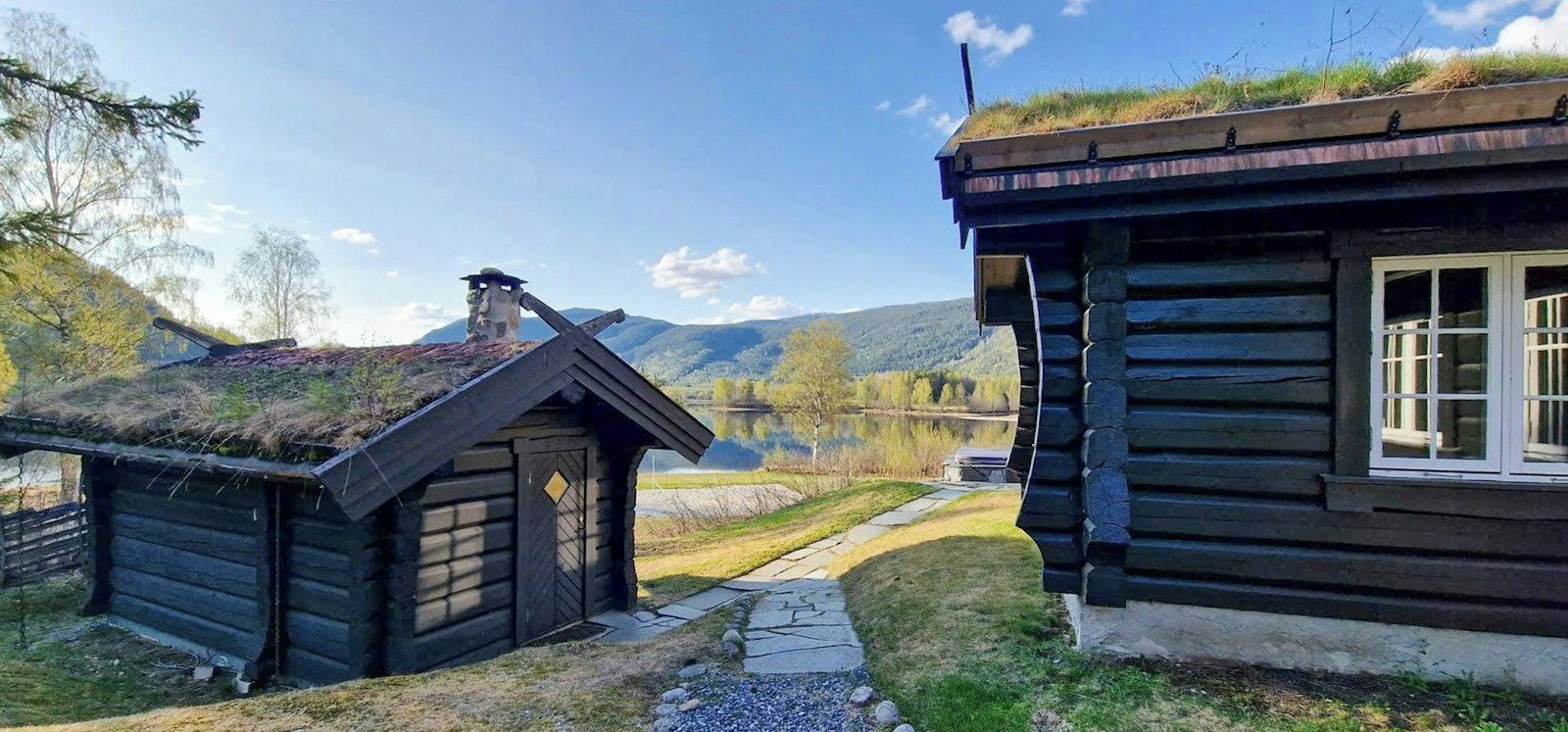Una cabaña de troncos con techo de paja, un sendero de piedra y vistas a un lago y montañas.