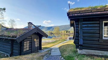 Una cabaña de troncos con techo de paja, un sendero de piedra y vistas a un lago y montañas.