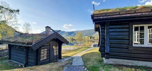 Hirsimökki, jossa on olkikatto, kivipolku ja näkymä järvelle ja vuorille.