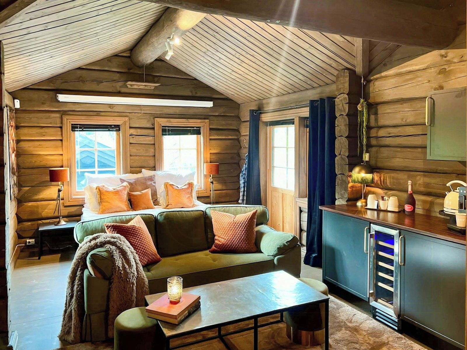Interior acogedor de una cabaña de troncos con un sofá verde, una mesa de comedor de madera y una zona de cocina con un refrigerador.