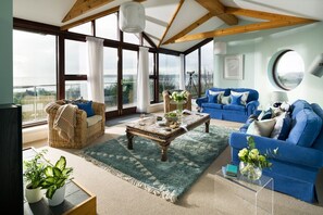 A spacious living room with a large window, a blue sofa set, a wooden coffee table, and a wicker chair. - Little Acres - sleeps 12 guests  in 5 bedrooms (Marazion)