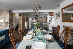 Uma sala de jantar com mesa posta para seis pessoas, rodeada por cadeiras de madeira, uma parede de pedra e uma cozinha ao fundo.