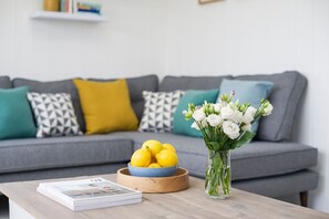 A living room with a grey sofa, a wooden coffee table, a bowl of lemons, and a vase of white flowers. - Skylark Beach House - sleeps 6 guests  in 3 bedrooms (Hayle)