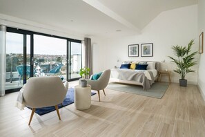 Une chambre moderne dotée d’un grand lit, d’un coin salon avec des fauteuils, d’une vue sur la ville grâce à de grandes fenêtres et d’un balcon aménagé avec du mobilier d’extérieur.
