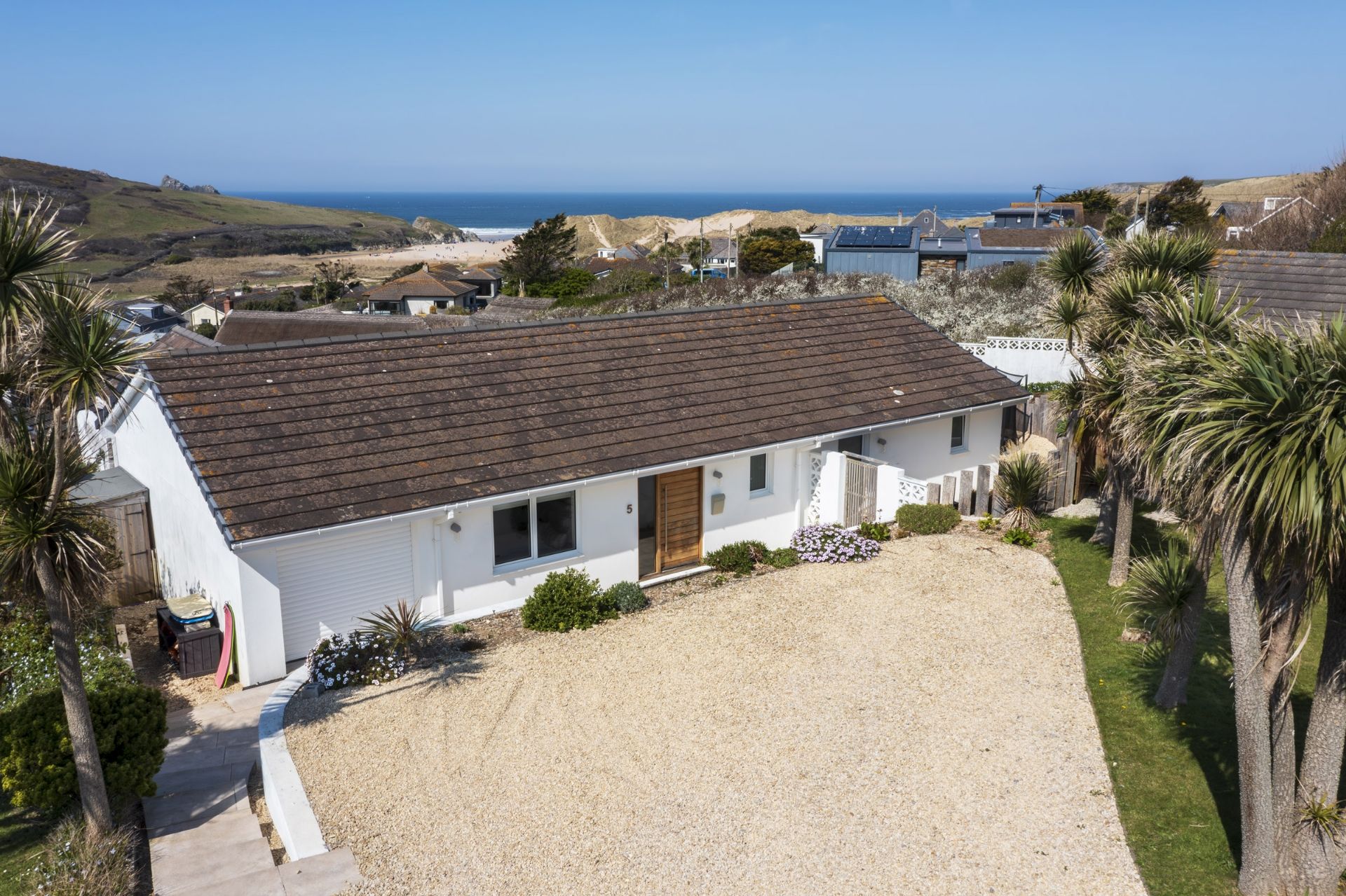 A white house with a gravel driveway and a wooden door, surrounded by palm trees and a view of the ocean.